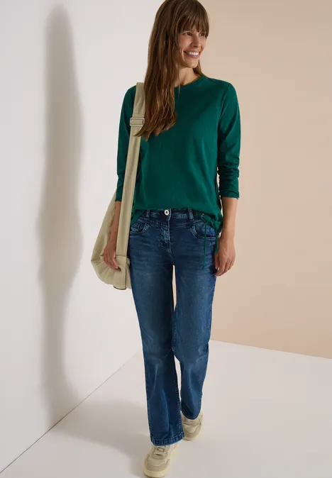 Langarm Shirt mit Rundhals und Tunnelzug dark fir green Eine Frau in grünem Pullover, blauen Jeans und Stofftasche lehnt lächelnd an einer Wand.