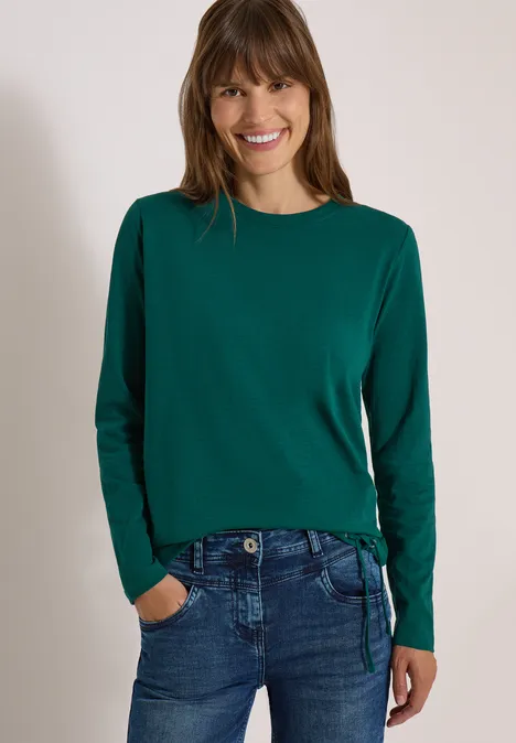 Langarm Shirt mit Rundhals und Tunnelzug dark fir green Frau in grünem Pullover und Jeans, lächelnd, Hände in den Taschen.