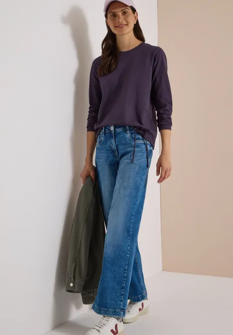 Langarm Shirt mit Rundhals und Tunnelzug lilac shadow Eine Frau lehnt an einer Wand und trägt ein violettes Longsleeve, weite Jeans, weiße Sneaker und eine rosa Cap.