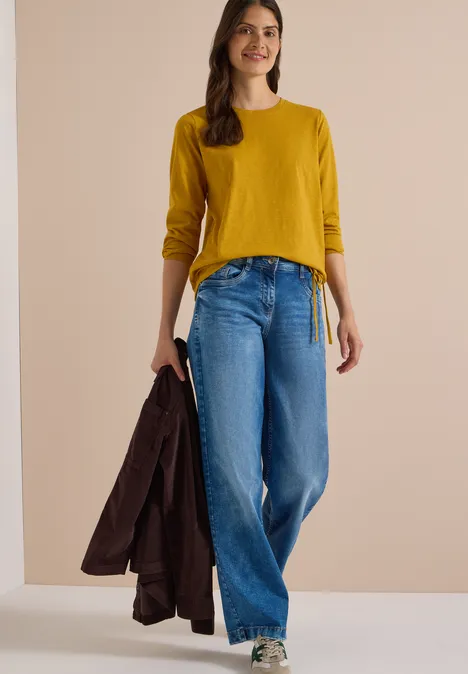 Langarm Shirt mit Rundhals und Tunnelzug golden honey Frau in senfgelbem Oberteil und weitem Jeans, die einen Mantel in der Hand hält.