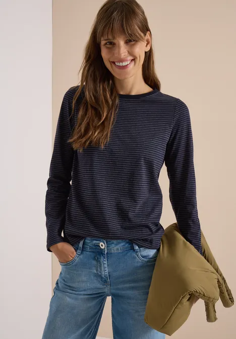 Femme en jean et pull rayé, tenant une veste, souriante.