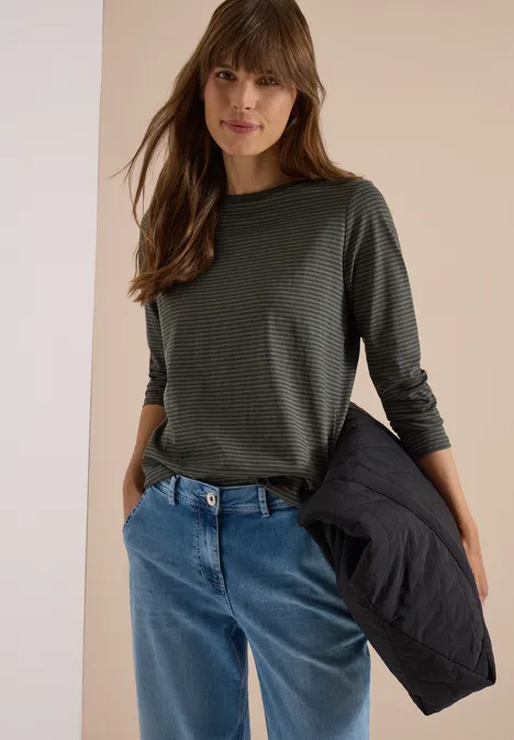 Une femme porte un t-shirt vert à manches longues et un jean bleu, tenant une veste noire.