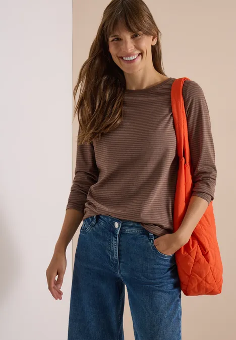Lächelnde Frau in braunem Langarmshirt, Jeans und oranger Umhängetasche.