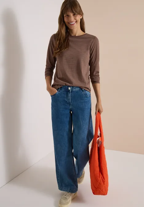 Frau in braunem Langarmshirt, weiten Jeans und weißen Sneakers hält eine orangefarbene Tasche und lächelt.