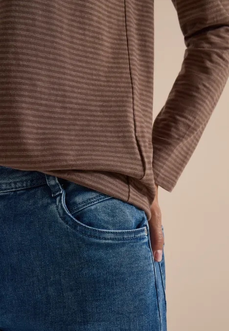 Person in braun gestreiftem Oberteil mit Hand in Jeans-Tasche vor beigem Hintergrund.