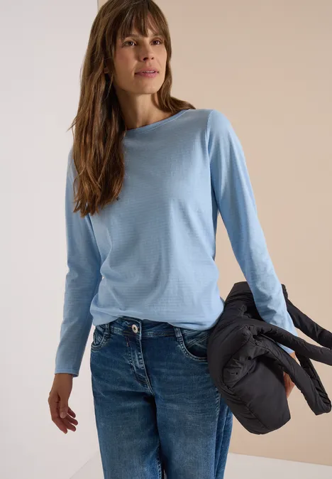Une femme en T-shirt à manches longues bleu clair tient une veste noire matelassée.
