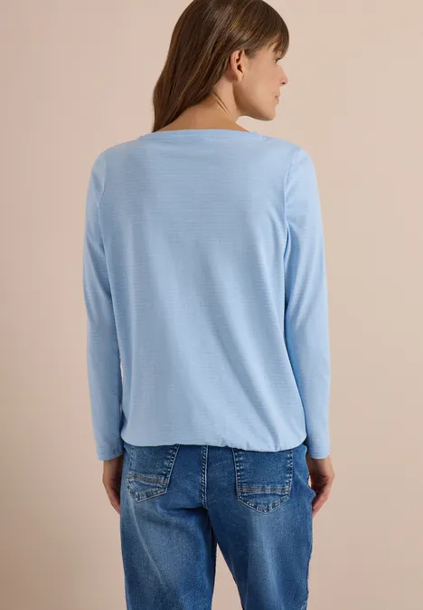 Femme de dos portant un haut bleu clair à manches longues et un jean sur fond neutre.