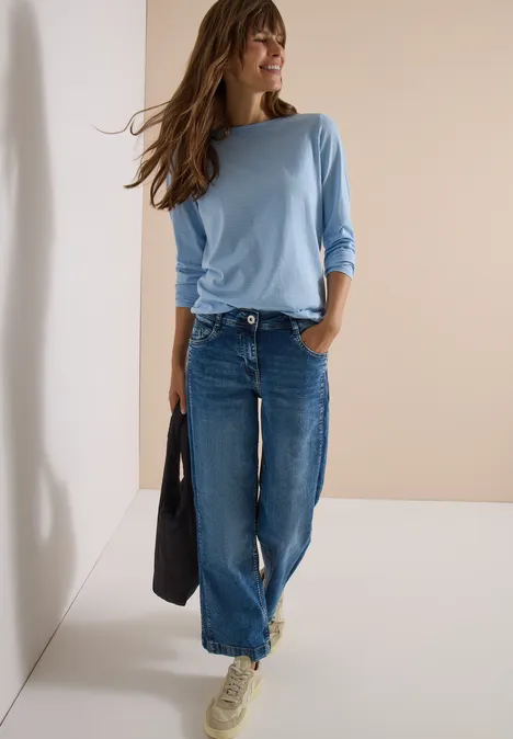 Femme en top manches longues bleu clair et jeans larges, sac noir, fond neutre.