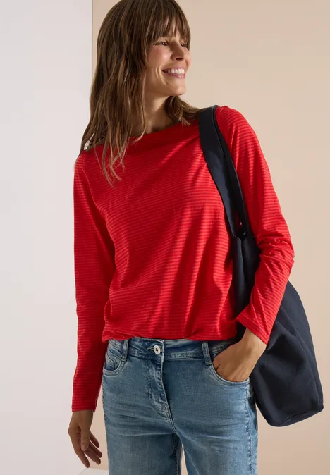 Femme souriante en chemise rouge à manches longues et jean, avec un sac épaule noir.