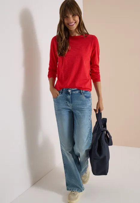 Femme souriante en pull rouge et jean ample, appuyée contre un mur blanc, tenant un sac bleu foncé.