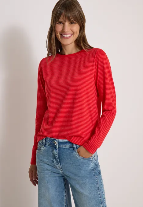 Femme souriante portant un t-shirt rouge à manches longues et un jean bleu sur fond blanc.