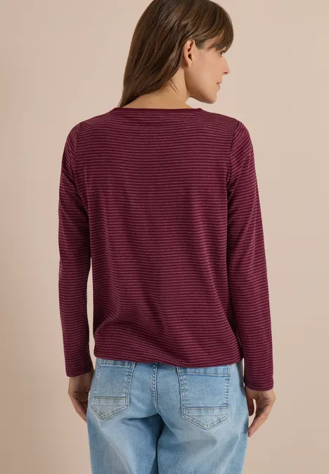 Vue de dos: femme en pull manches longues bordeaux à fines rayures et jean bleu devant un mur beige.