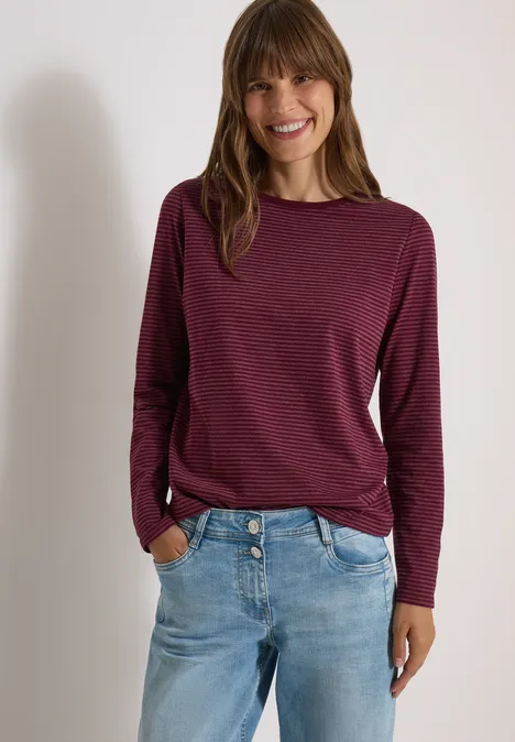 Femme en pull bordeaux à manches longues et jean, souriante, mains dans les poches.