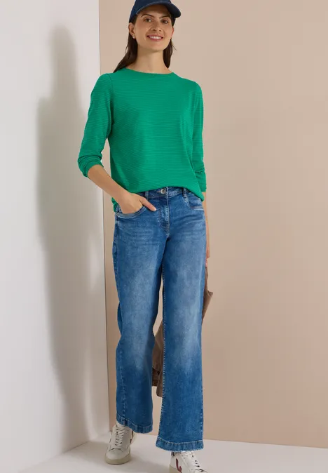 Eine Frau in grünem Pullover, weiter blauer Jeans, weißen Sneakers und Mütze vor hellem Hintergrund.