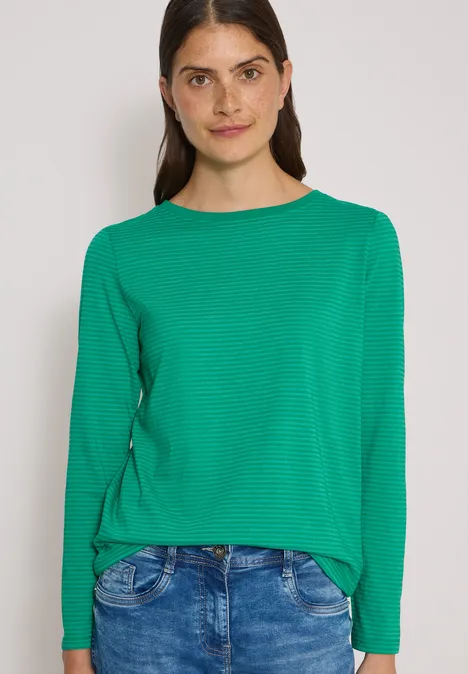 Eine Frau mit langärmligem grünen Shirt und Jeans vor weißem Hintergrund lächelnd.