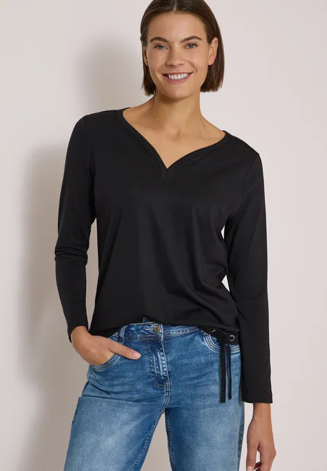 Shirt mit Split Neck im Tunika-Look Black Eine Frau mit schwarzem Langarmshirt und blauen Jeans, lächelnd vor neutralem Hintergrund.
