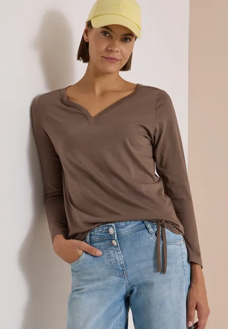 Shirt mit Split Neck im Tunika-Look urban taupe Frau trägt braunes V-Neck-Shirt mit seitlichem Kordelzug, blaue Jeans und gelbe Cap vor neutralem Hintergrund.