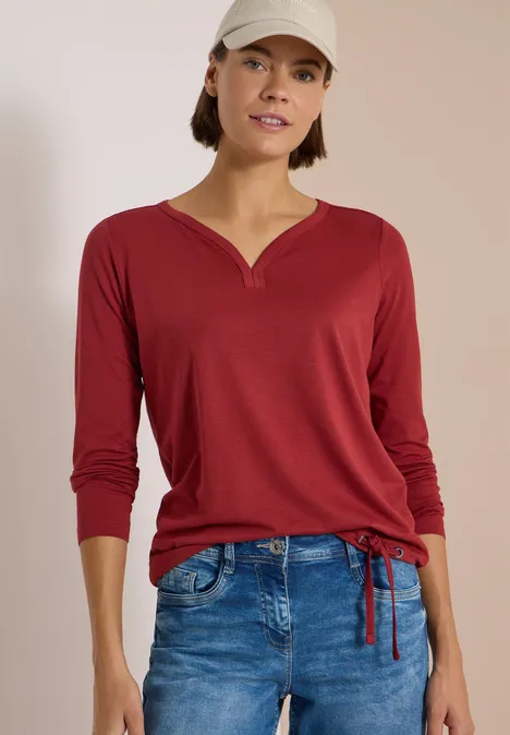 Shirt mit Split Neck im Tunika-Look russet red Frau in rotem Langarmshirt mit Schnürung am Saum, blauen Jeans und weißer Kappe vor hellem Hintergrund.