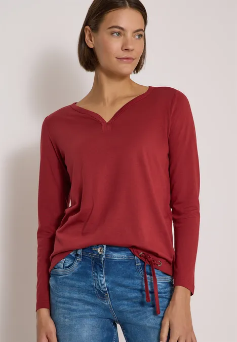 Shirt mit Split Neck im Tunika-Look russet red Frau mit rotem Langarmshirt und blauen Jeans steht vor hellem Hintergrund.