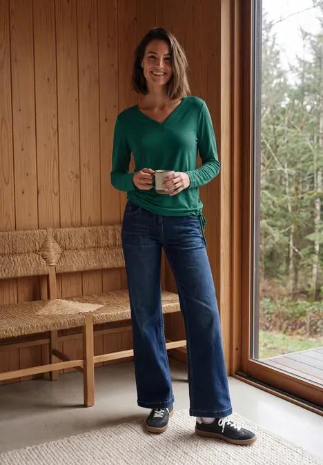 Een vrouw in een groen bloesje en spijkerbroek, glimlachend met een mok in een lichte houten ruimte bij een raam.