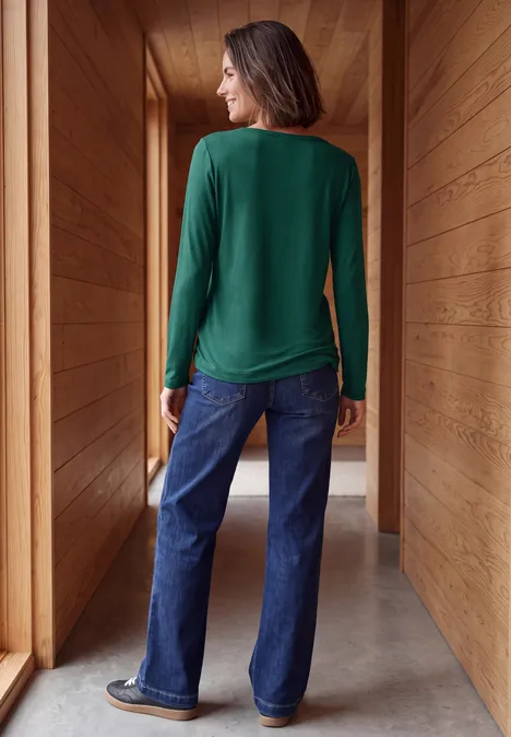 Vrouw in groen shirt en wijde jeans in een houten gang, kijkt opzij.