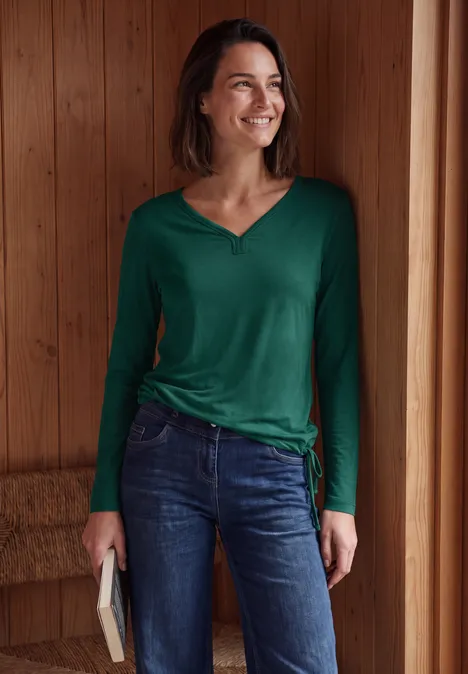 Een lachende vrouw in groene top en spijkerbroek staat in een houten ruimte en houdt een boek vast.