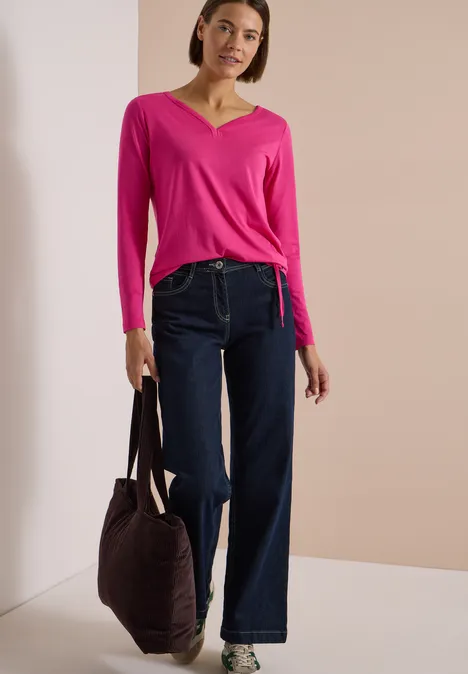 Shirt mit Split Neck im Tunika-Look pink flash Frau trägt pinken V-Ausschnitt-Pullover, weite Jeans-Hose und hält Tasche vor beigem Hintergrund.