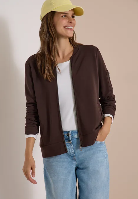 Ottoman Blouson in Unifarbe truffle brown Eine Frau in lässiger Kleidung: brauner Hoodie, weißes T-Shirt, blaue Jeans und gelbe Kappe, lächelnd.