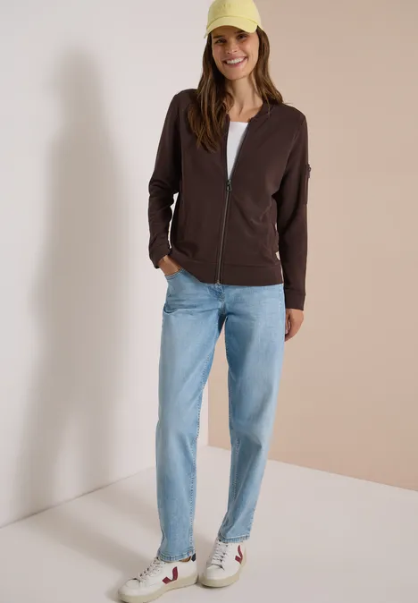 Ottoman Blouson in Unifarbe truffle brown Frau in Jeans, weißem Shirt, brauner Jacke, weißen Sneakern und hellgrünem Cap vor neutralem Hintergrund.