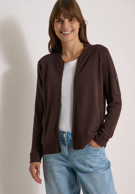 Ottoman Blouson in Unifarbe truffle brown Lächelnde Frau in brauner Bomberjacke über weißem Shirt und Jeans vor hellem Hintergrund.