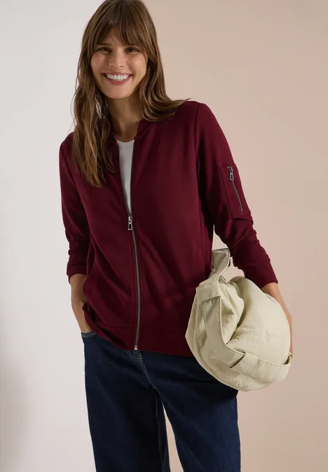 Ottoman Blouson in Unifarbe red wine Fröhliche Frau in burgunderroter Jacke, weißem Shirt und Jeans hält eine beige Tasche.