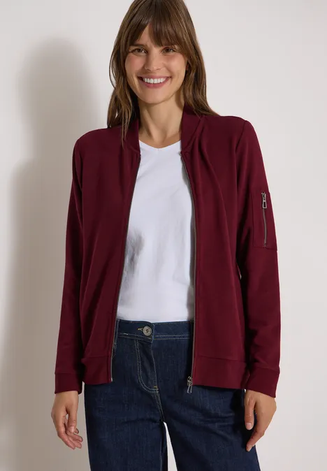 Ottoman Blouson in Unifarbe red wine Eine lächelnde Frau trägt eine bordeauxrote Jacke, weißes T-Shirt und blaue Jeans vor hellem Hintergrund.