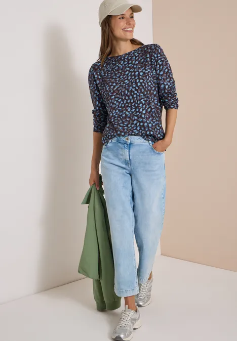 Femme en jean, chemisier à motifs et casquette tient une veste verte