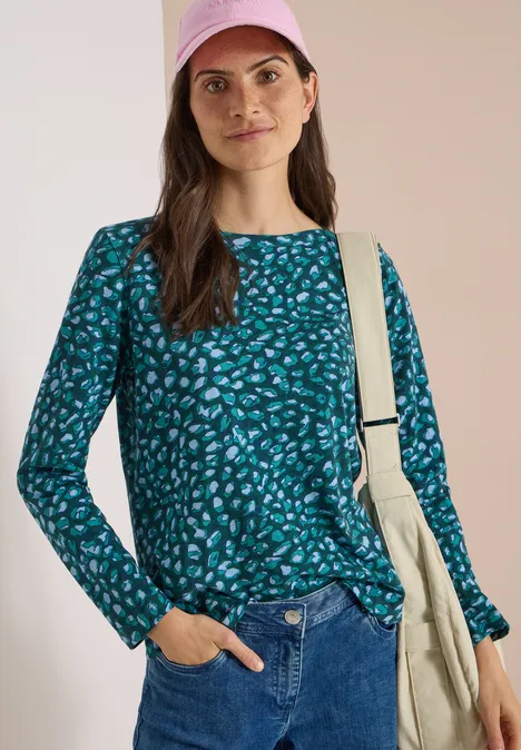 Femme en tee-shirt vert à motifs et jean, avec sac sur l'épaule, posant devant un fond clair.