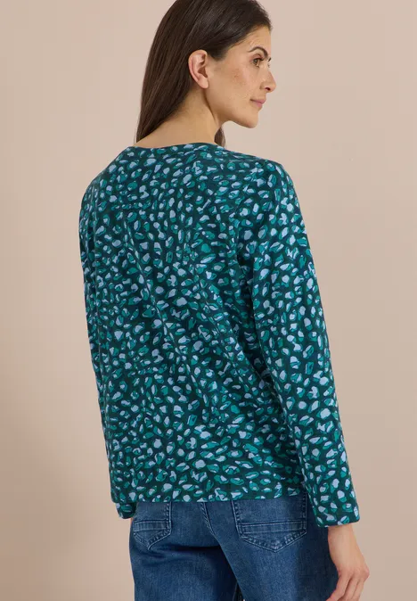 Vue de dos, une femme aux cheveux bruns porte un haut bleu-vert à motif et un jean.