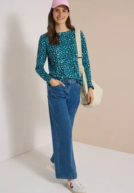 Femme souriante en pull bleu à pois, jean large, casquette rose et sac blanc devant un mur clair.