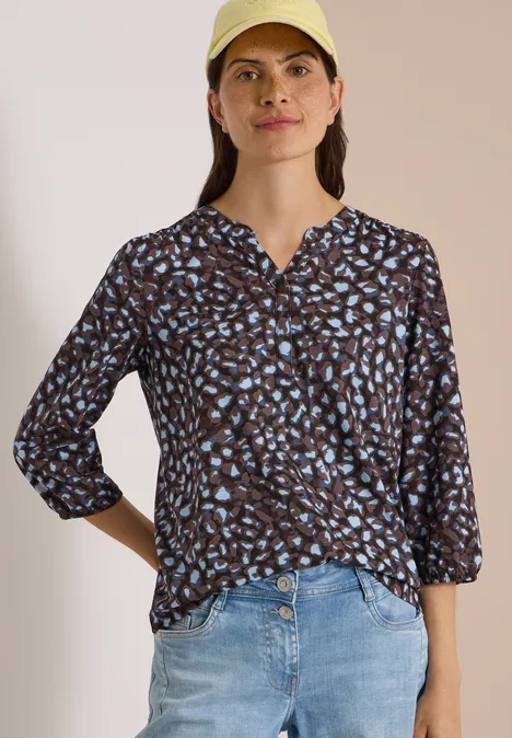 Femme en casquette jaune porte une blouse noire à motif feuilles bleu-blanc et un jean clair.