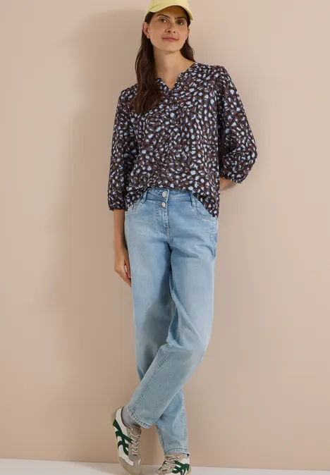 Femme en blouse à motifs, jean ample, baskets et casquette jaune devant fond beige