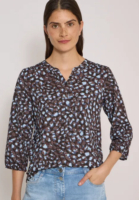 Femme portant une blouse à motif floral bleu et un jean, fond clair.