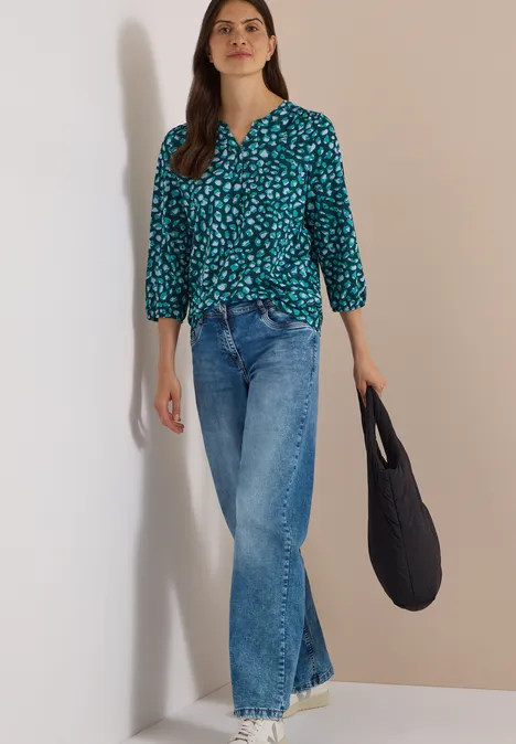 Femme en chemisier vert à motifs, jean bleu et sac noir devant un fond neutre.