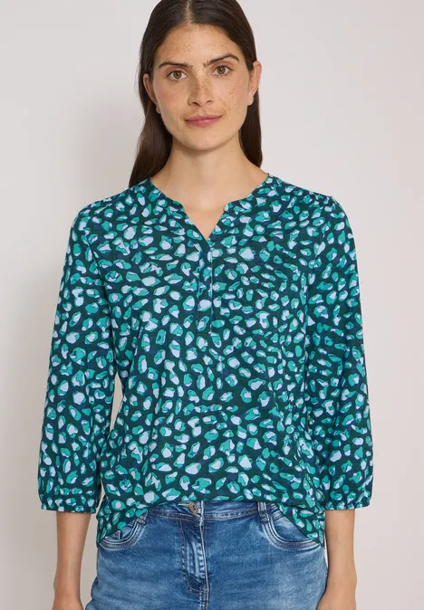 Jeune femme en chemisier turquoise à motifs et manches 3/4 avec jean, cheveux lâchés, léger sourire.