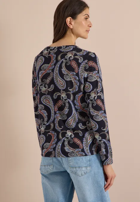 Frau von hinten in Jeans und dunkelblauem Paisley-Oberteil vor hellem Hintergrund.