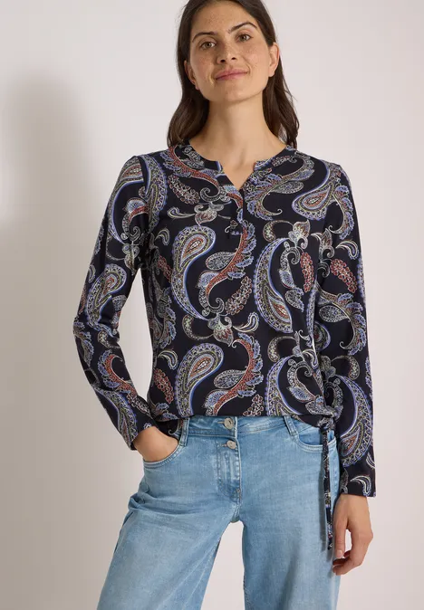 Frau in hellblauen Jeans und dunkelblauer, paisley-bedruckter Bluse mit Knoten am Saum, lächelnd.