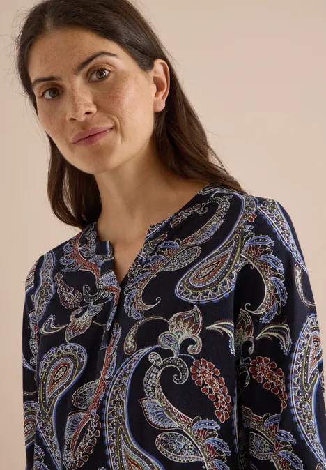 Frau mit dunklen Haaren in dunkelblauer Bluse mit buntem Paisley-Muster vor hellem Hintergrund.