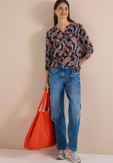 Frau trägt Paisley-Bluse, weite Jeans und hält eine große orangene Tasche vor beigem Hintergrund.