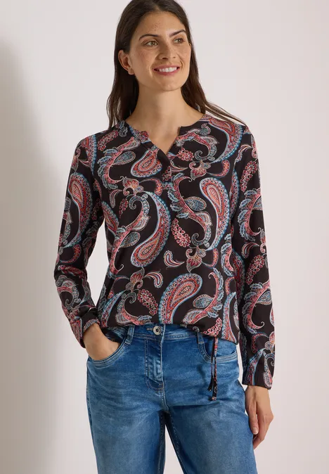 Eine Frau trägt eine langärmlige Bluse mit Paisley-Muster und Jeans.