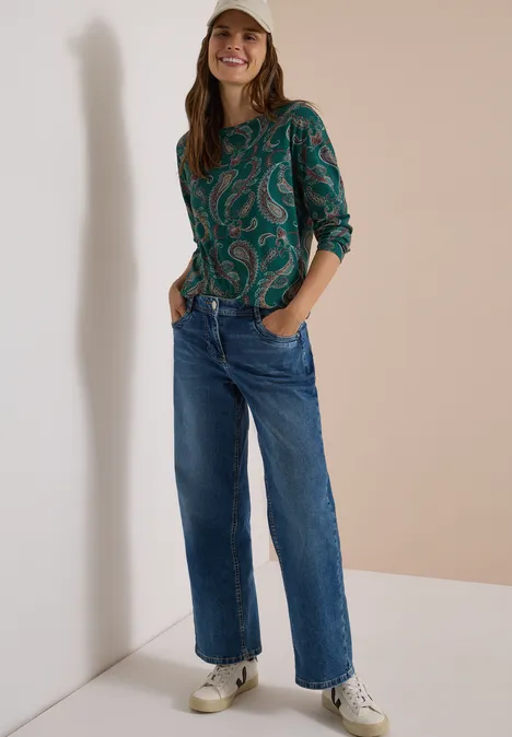 Eine junge Frau trägt Jeans, weiße Sneaker und eine grüne gemusterte Bluse, sie lächelt in neutralem Umfeld.