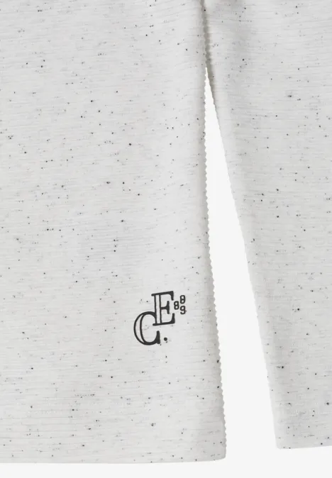 Zbliżenie kremowego melanżowego swetra z małym czarnym logo przy dolnym brzegu.