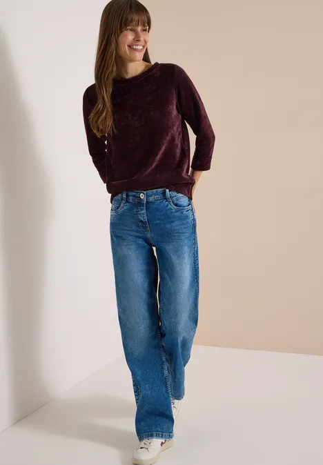 Frau in weitem blauen Jeans und burgunderfarbenem Pullover, die lächelnd einen hellen Raum betritt.
