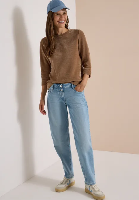 Vrouw leunt tegen muur, draagt bruin trui, lichtblauwe jeans, witte sneakers en blauwe pet.